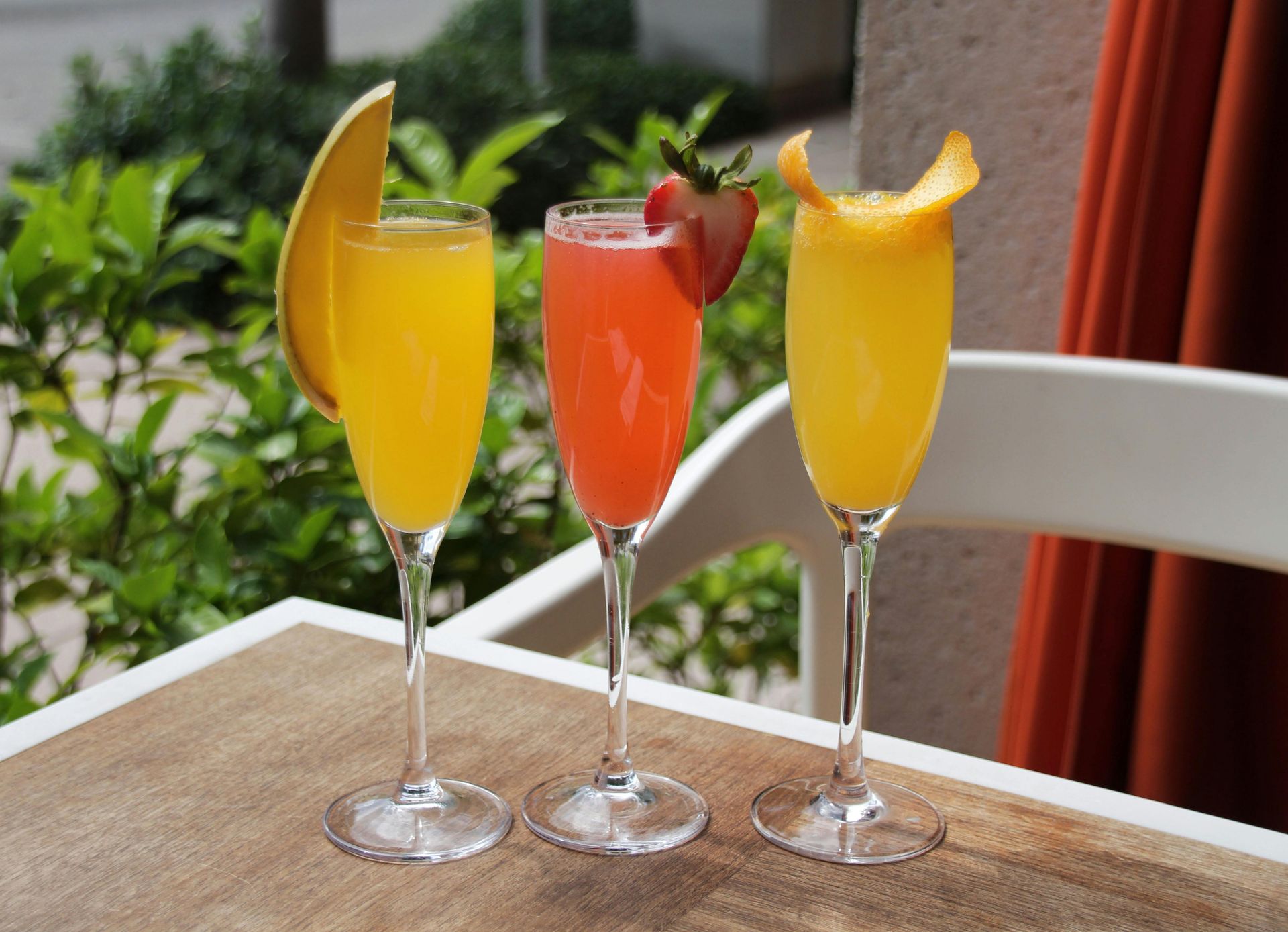 Three champagne flutes with colorful cocktails, garnished with fruit, on a table.