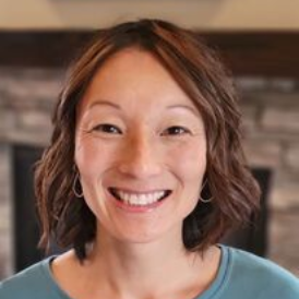 Woman smiling, wearing a blue shirt. Brown hair, circular earrings. Blurred stone fireplace background.