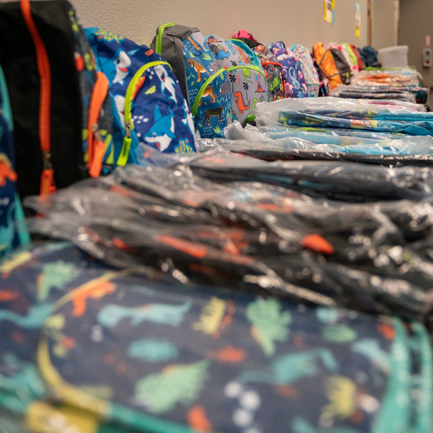 Backpacks in various patterns and colors stacked on a table, ready for distribution.