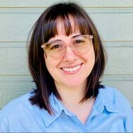 Woman with short dark hair, bangs, and glasses smiles, wearing a blue shirt, in front of a green wall.