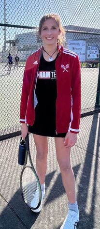 Woman in tennis attire holding a racket and water bottle on a tennis court, smiling.