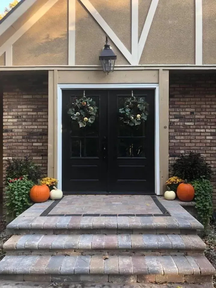 Black double doors with wreaths, flanked by pumpkins and greenery on brick steps.
