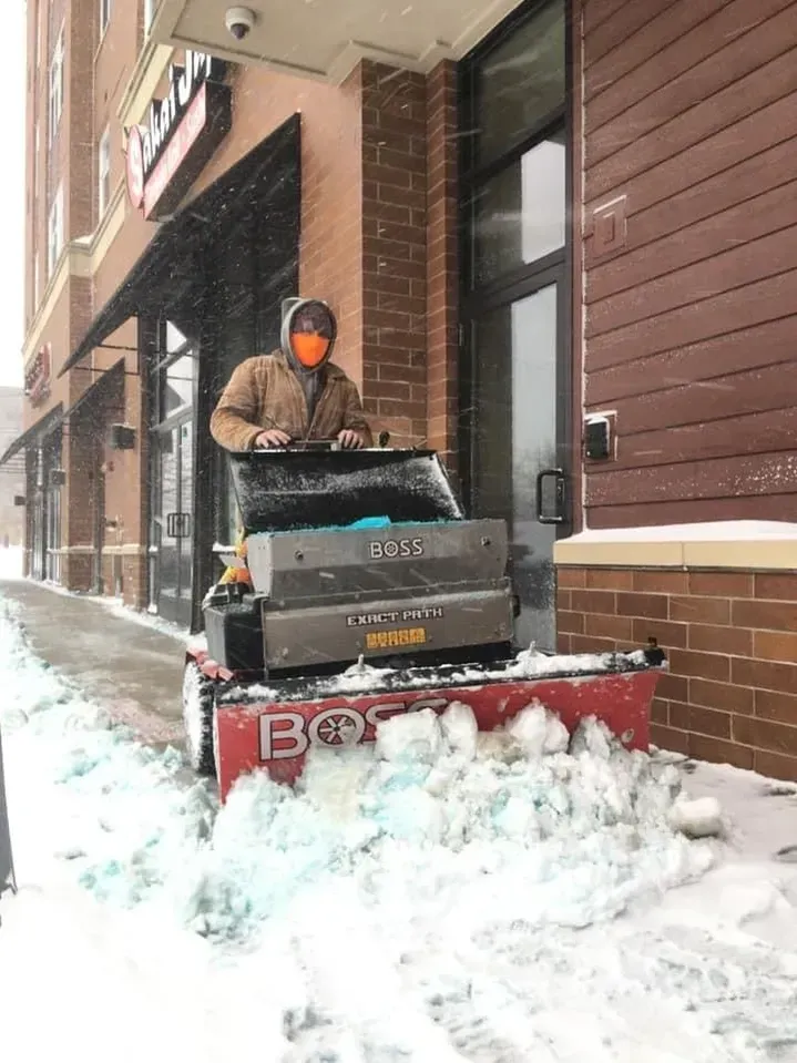 Person operating a snowblower on a snowy sidewalk outside a brick building.