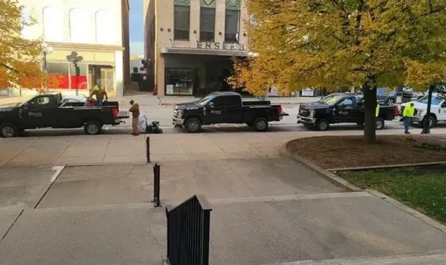 Several black pickup trucks parked on a street near buildings. People in reflective vests are visible.