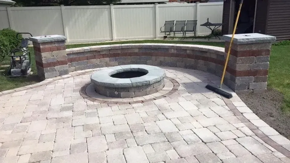 Stone fire pit and curved wall in a paved patio, near green grass, against a white fence.
