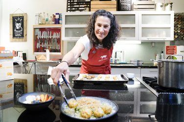 Woman in red apron plating food with tongs in a kitchen, smiling at the camera.