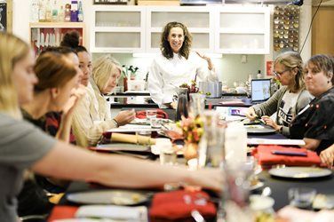 Group of people at a table, with a woman standing and gesturing, in a kitchen setting.