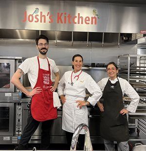 Three people pose in a kitchen, under a sign that reads 