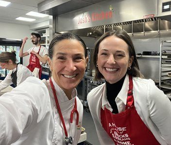 Two women smiling in a kitchen, one wearing a chef's coat and stethoscope, the other wearing a white shirt and red apron.