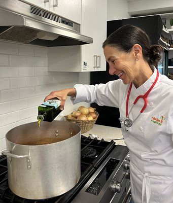 Woman in chef coat, with stethoscope, pouring oil into pot on stove. Smiling in kitchen.