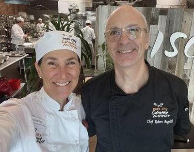 Two chefs smiling in a kitchen; one in a toque and white coat, the other in black.
