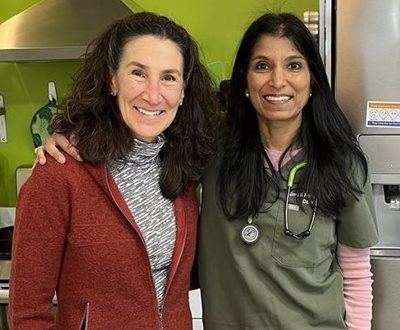 Two smiling women pose indoors. One wears a red jacket, the other a green medical scrub with a stethoscope.