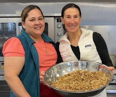 Two women holding a bowl of food, smiling in a kitchen. One wears a vest.
