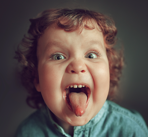 Child with curly red hair sticking out tongue, making a silly face with wide eyes.
