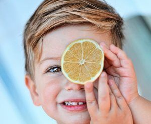 Child holding a lemon slice over one eye, smiling against a light blue background