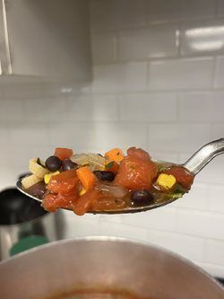 Spoonful of soup with vegetables and beans, held up in front of a white tiled wall.
