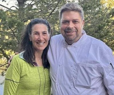 Woman in green top and chef in white coat smiling outside.