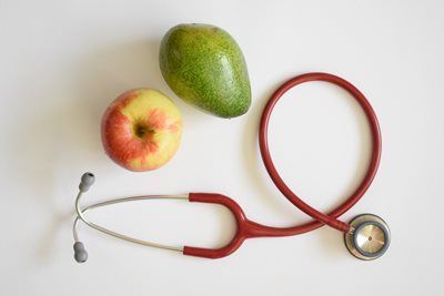 Red stethoscope next to an apple and avocado on a white surface.