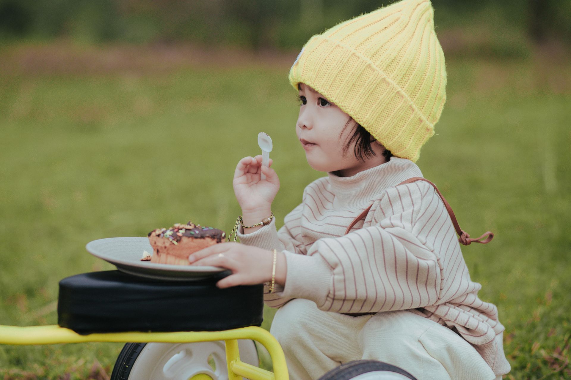 Child in yellow hat eats dessert outdoors.
