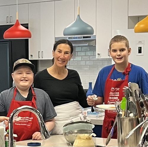 Three people in a kitchen, wearing aprons. One woman smiles between two smiling boys, with colorful pendant lights above.