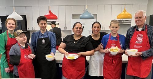 Group of people in red aprons in a kitchen, holding bowls of food. Overhead colored pendant lights.