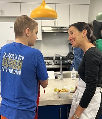 Person in blue shirt conversing with person in black shirt in a kitchen setting. They stand near a counter with food.