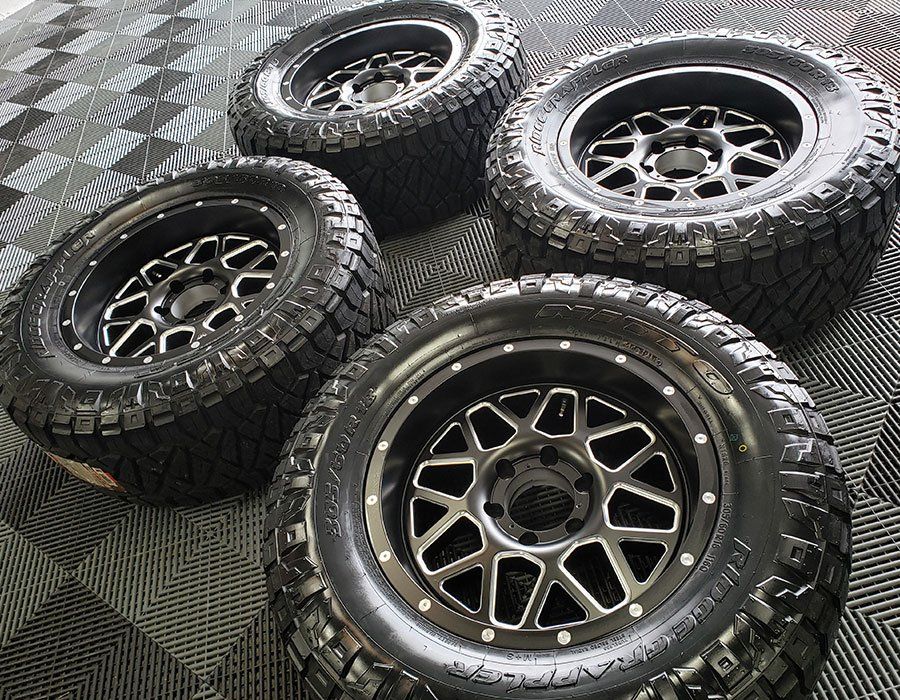 Four black off-road tires with silver rims, arranged on a textured floor.