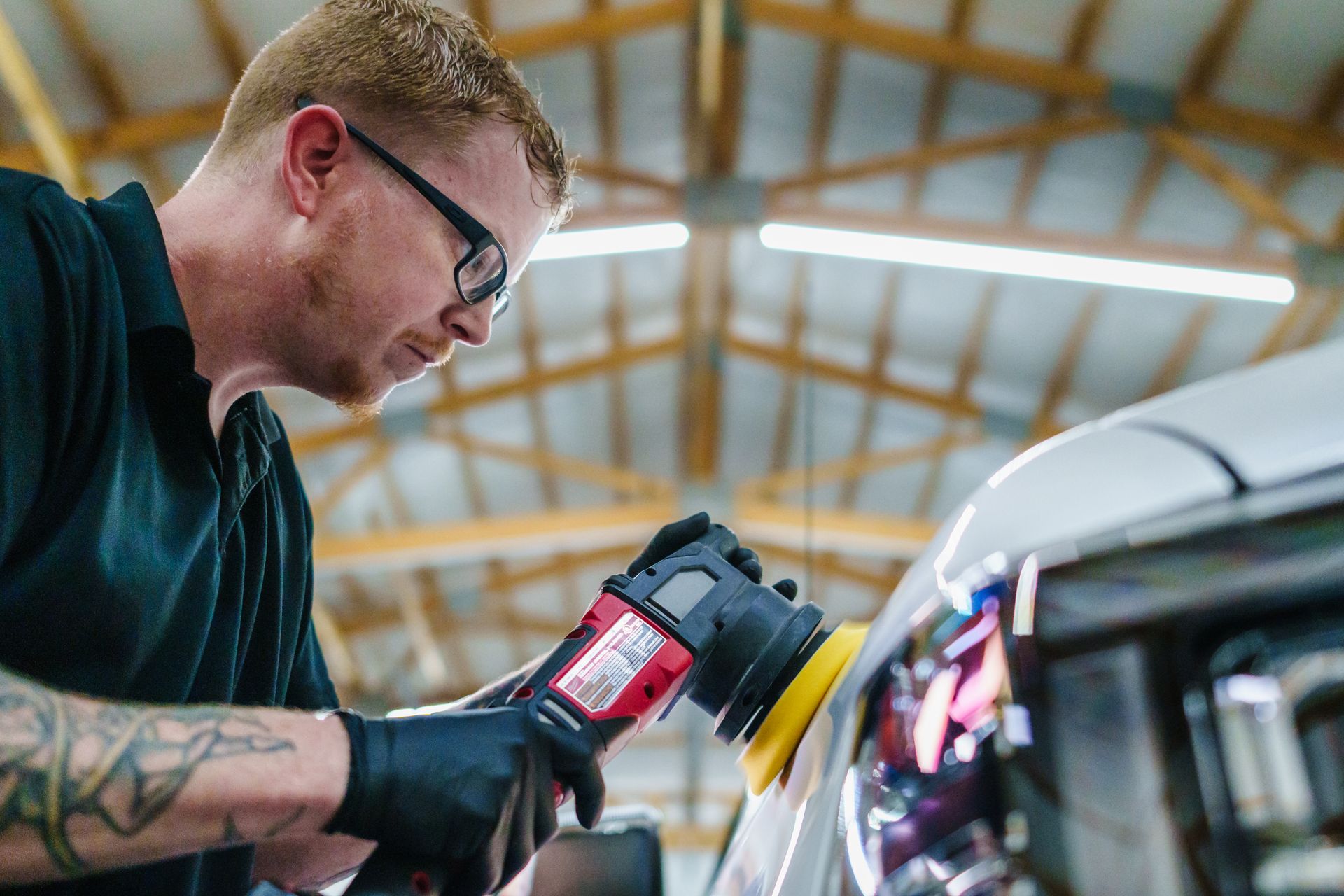 Man polishing a car's front panel, wearing glasses and gloves, in a well-lit garage.