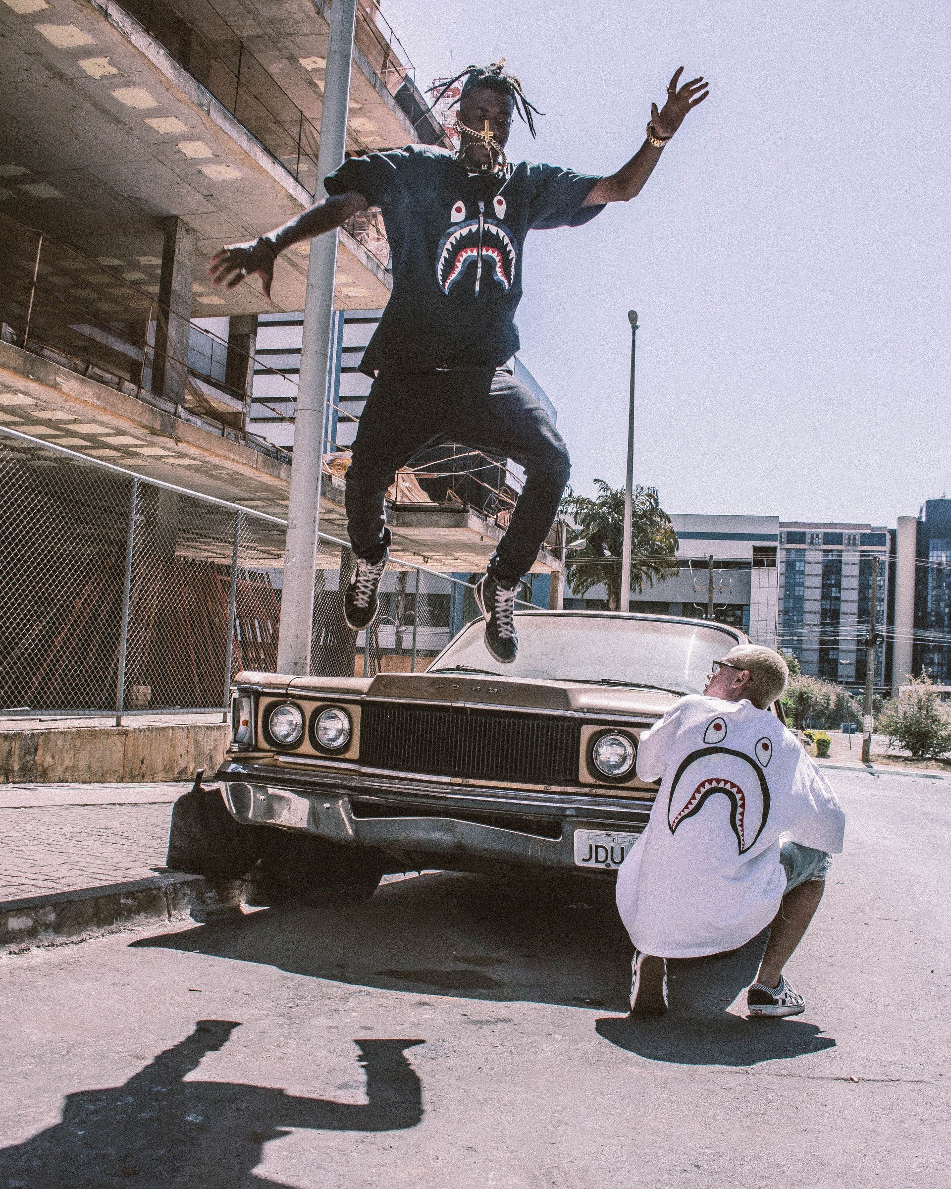 A man is jumping over a car while another man squats next to it.