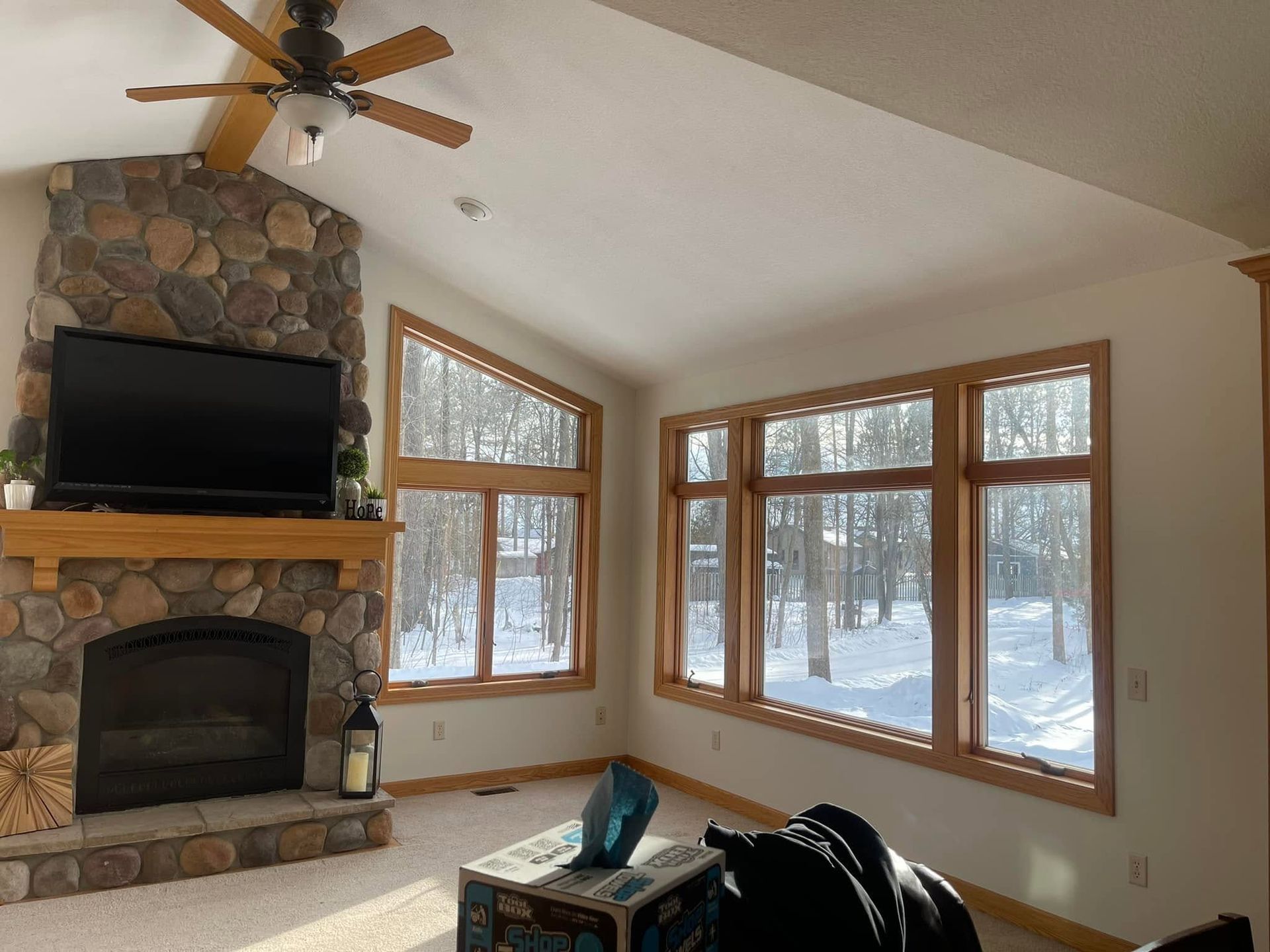 Living room with stone fireplace, large windows, ceiling fan, and snowy woods outside.