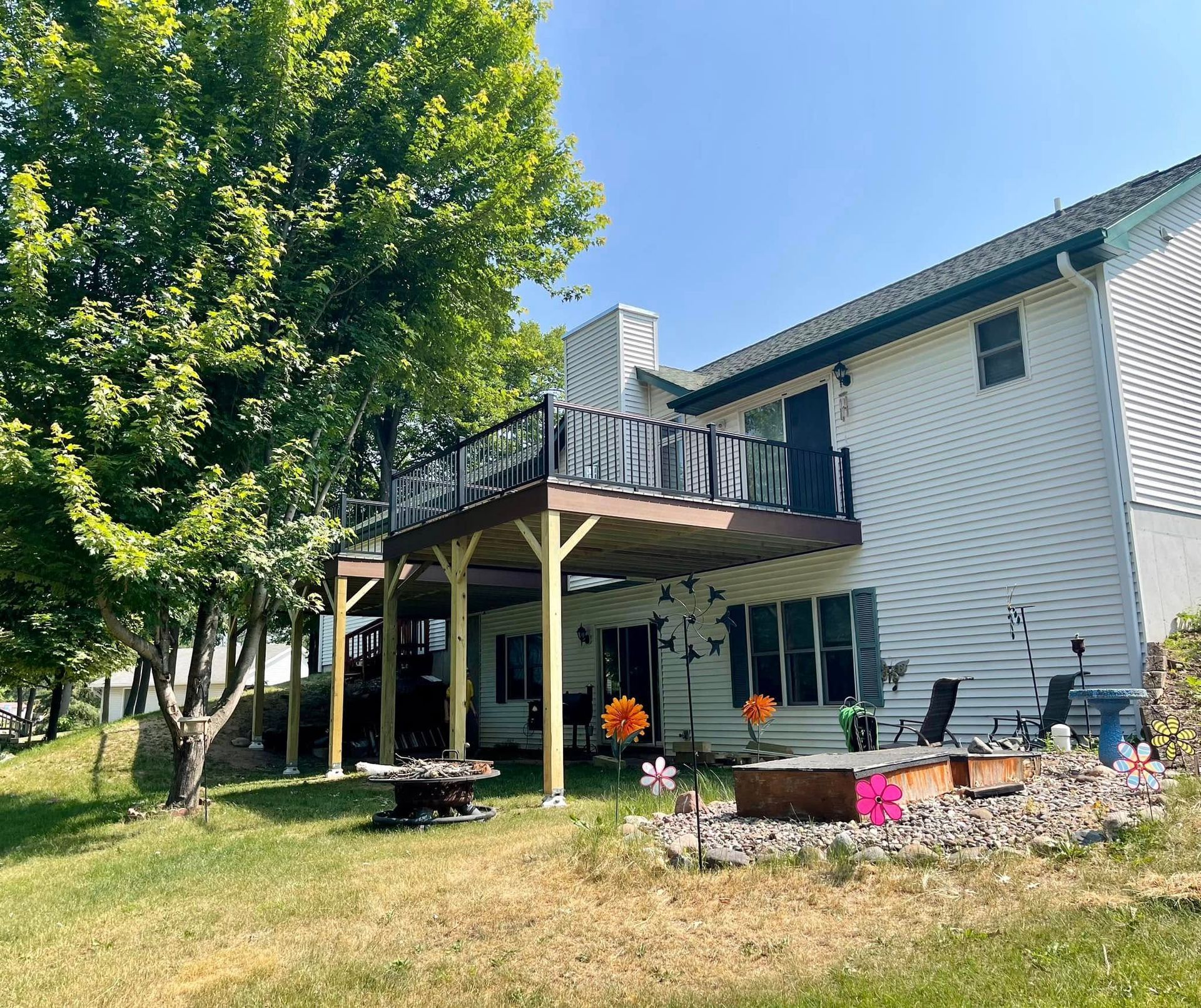 Two-story house with a wooden deck, patio furniture, and flower beds under a leafy tree on a sunny day