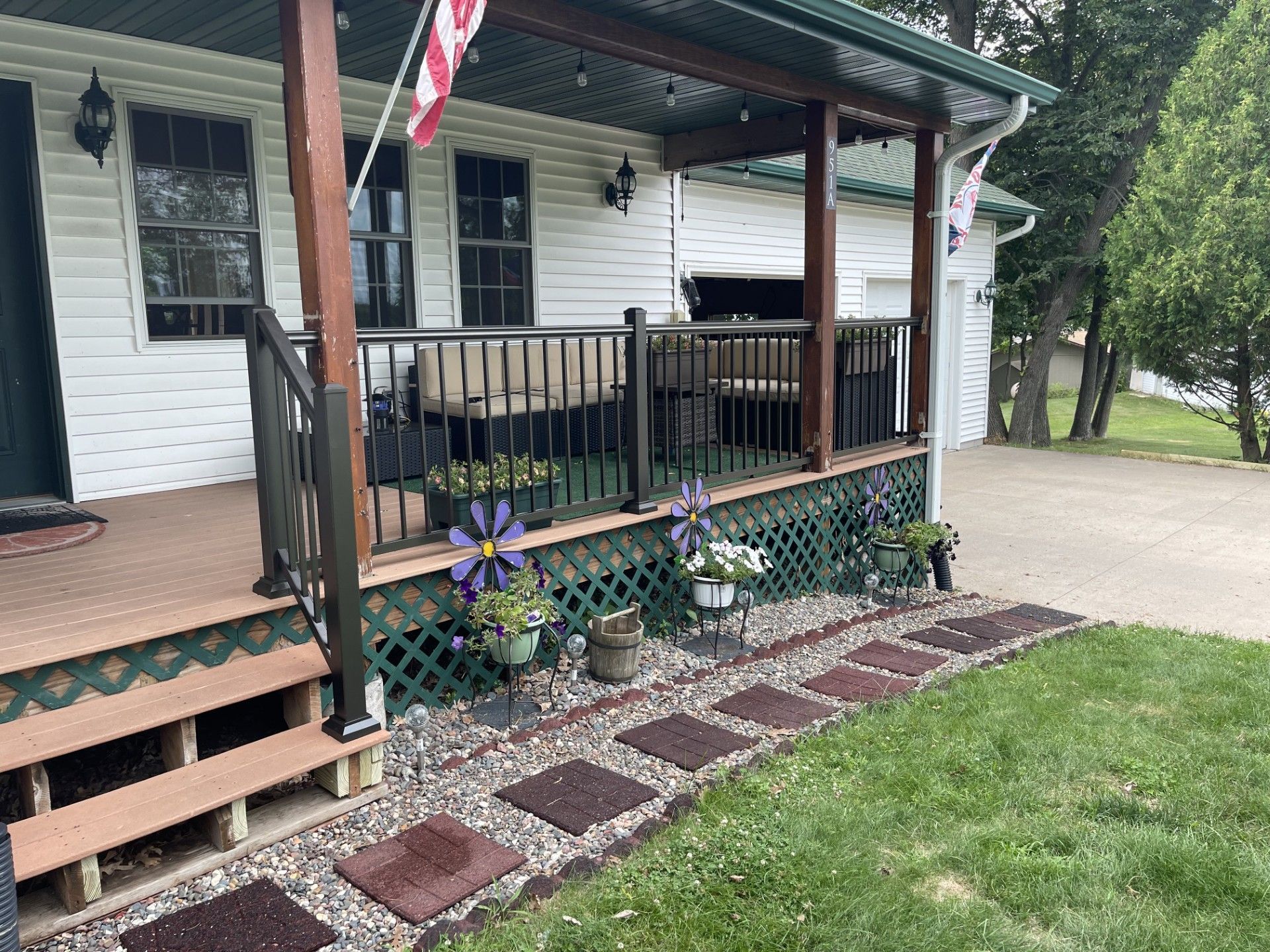 Front porch with green railing, potted flowers, and a brick walkway beside a white house