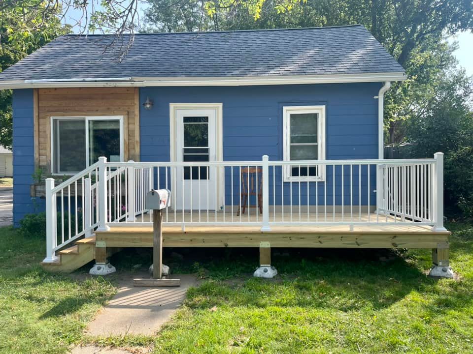 Blue house with a small wooden front deck and white railing in a grassy yard