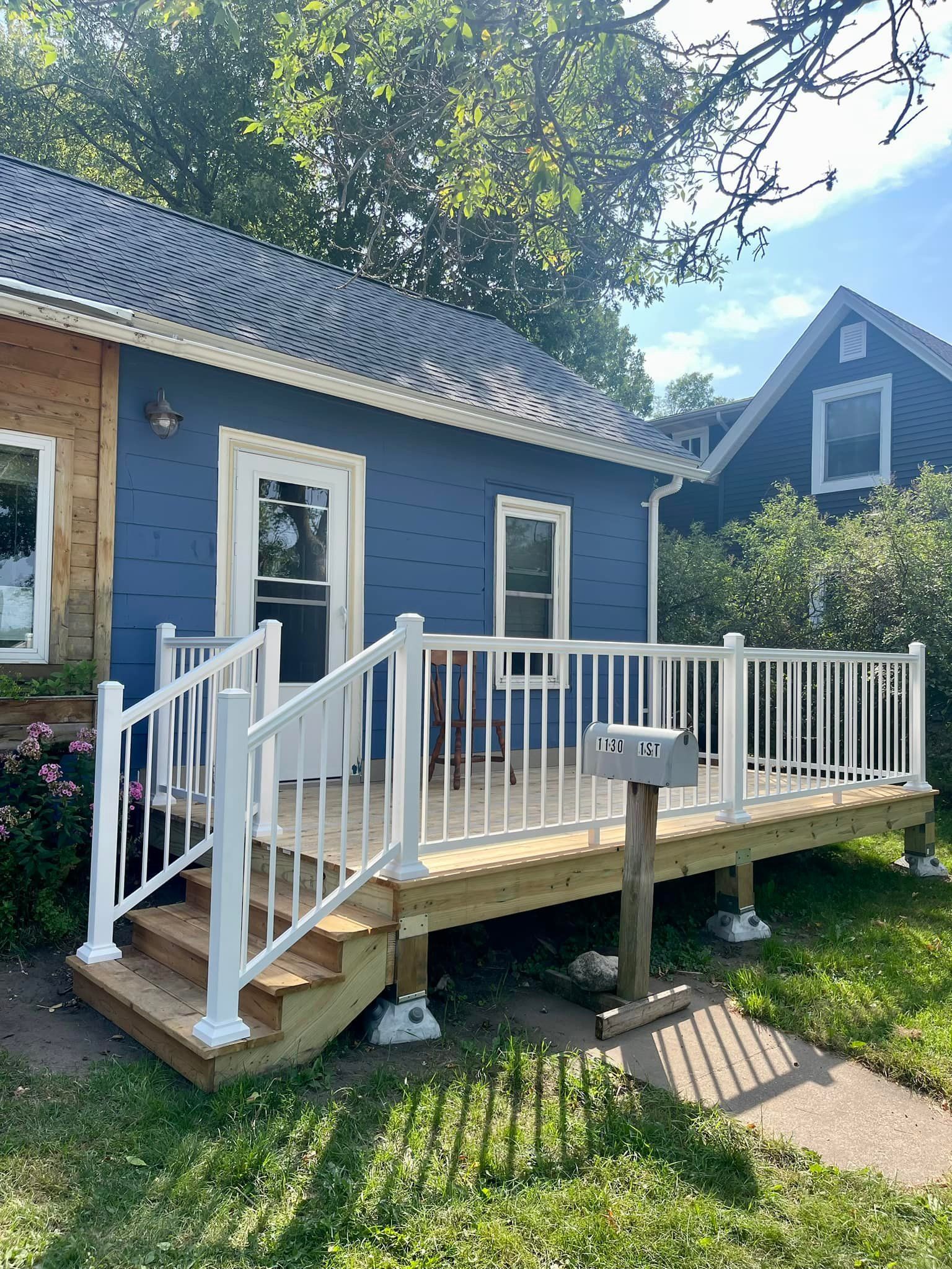 Blue house with a white-railed wooden porch, steps, and a mailbox in a sunny yard.