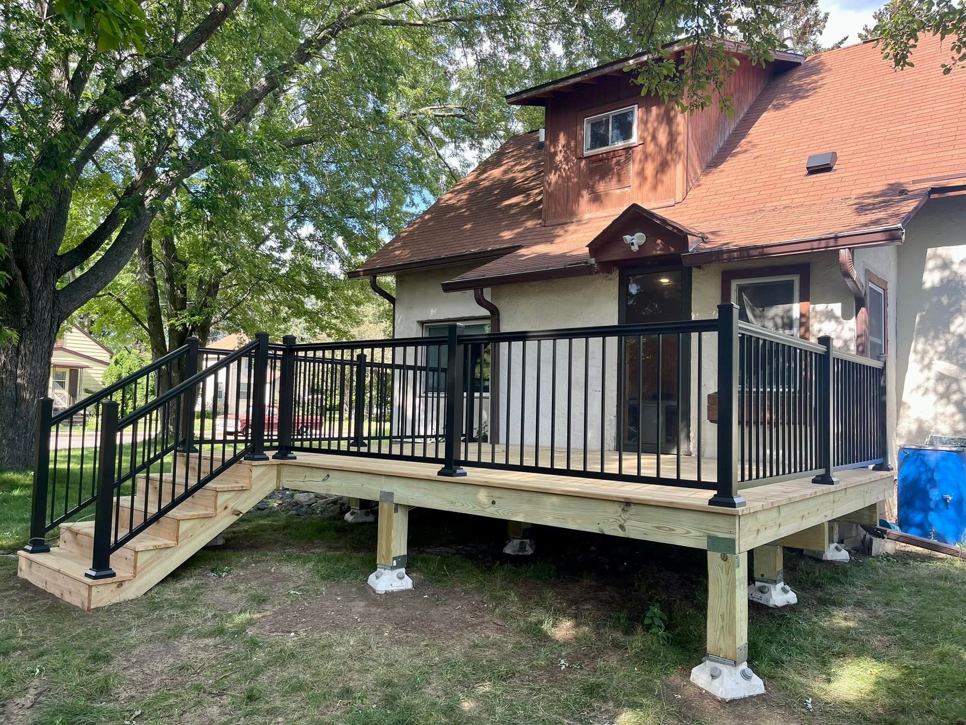 Raised wooden deck with black railings beside a house and trees in a yard.