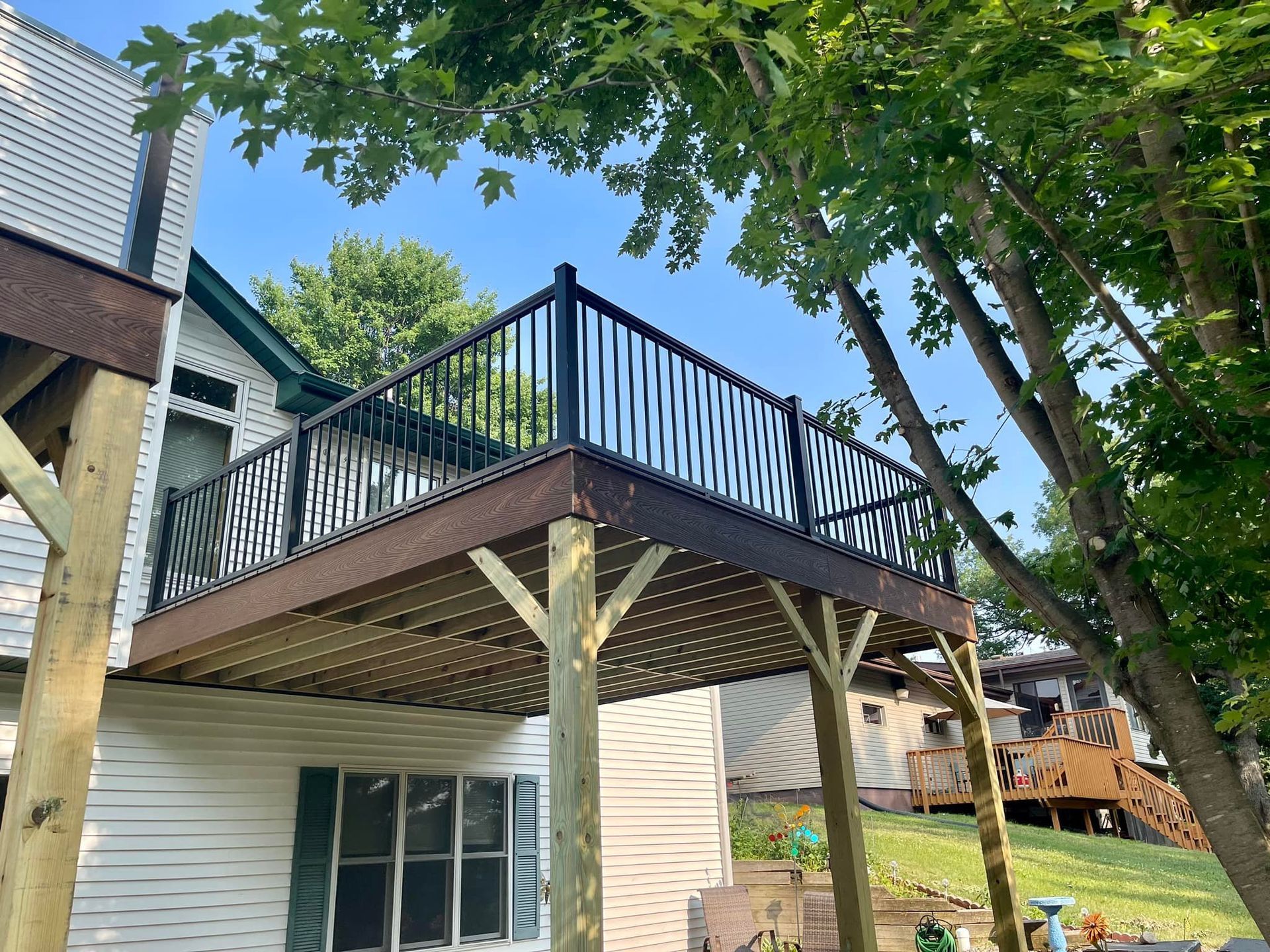 Back deck with black railing over a house, surrounded by trees and blue sky