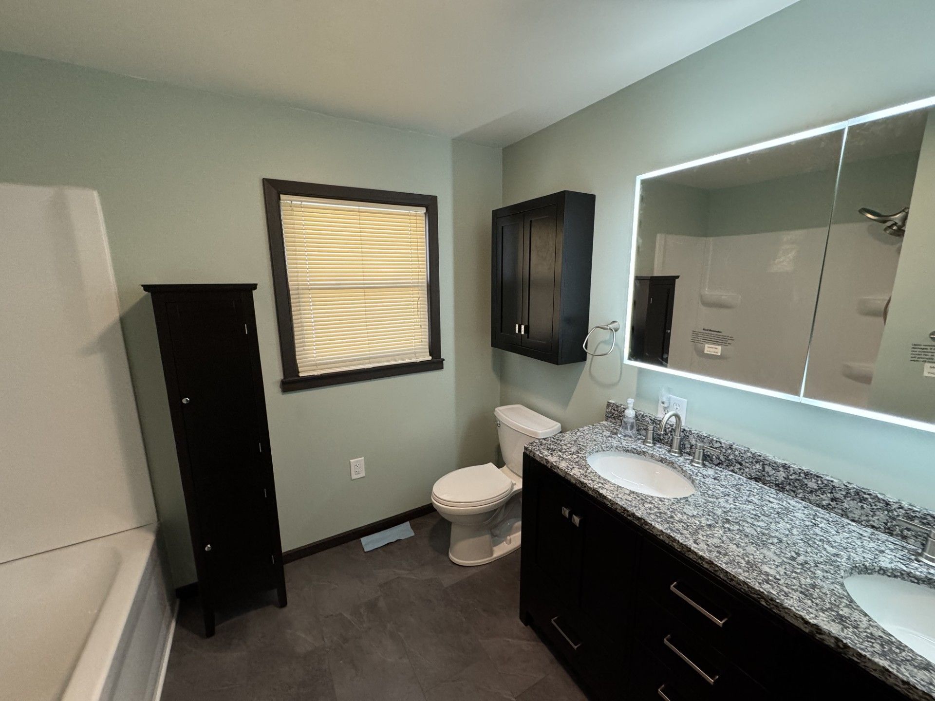 Modern bathroom with dark vanity, large mirror, toilet, window, and black cabinet.