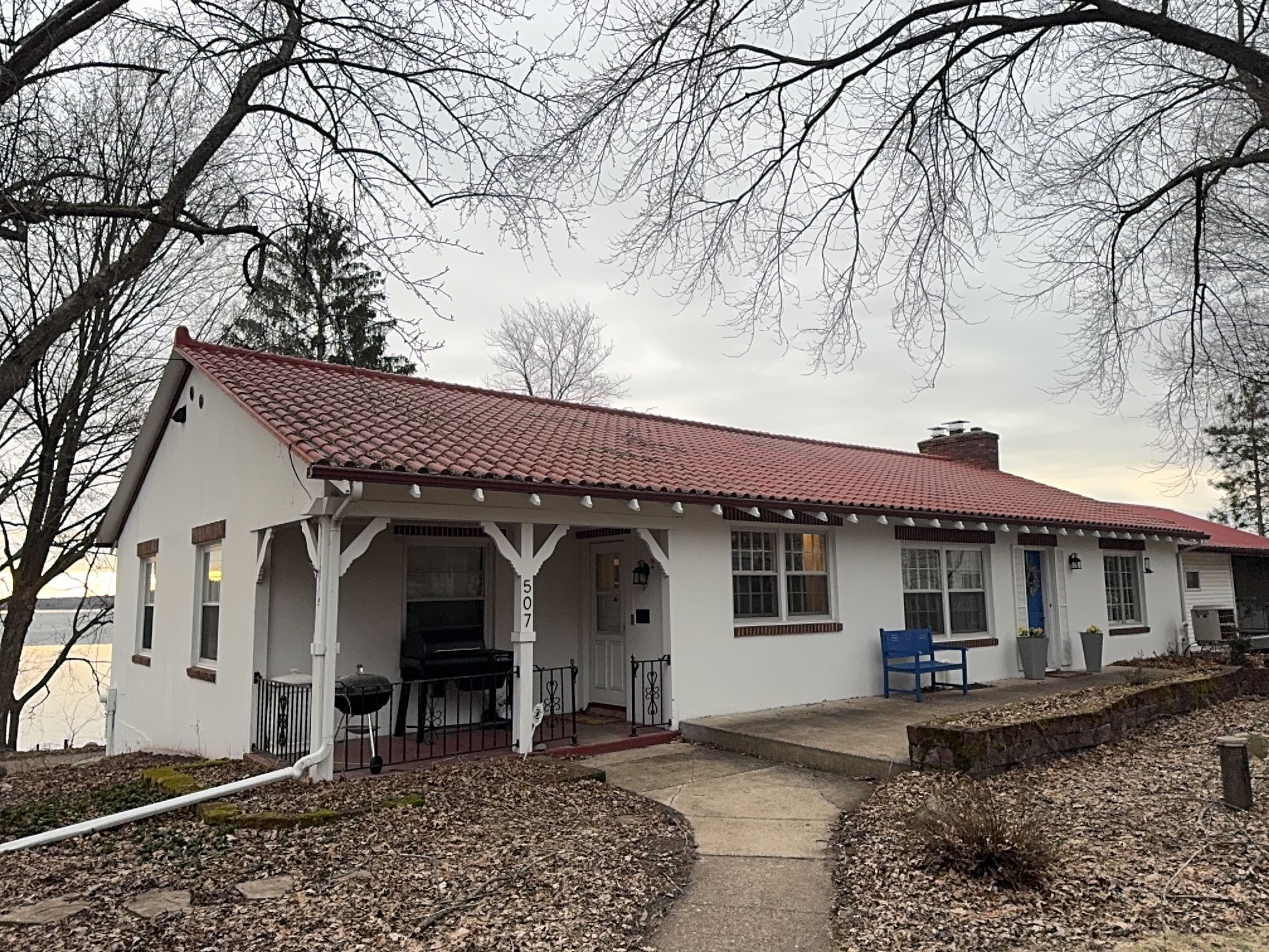 White house with red roof and porch, set among bare trees on a leaf-covered path.