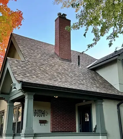 Small brick house with a steep shingled roof, tall chimney, and autumn trees overhead