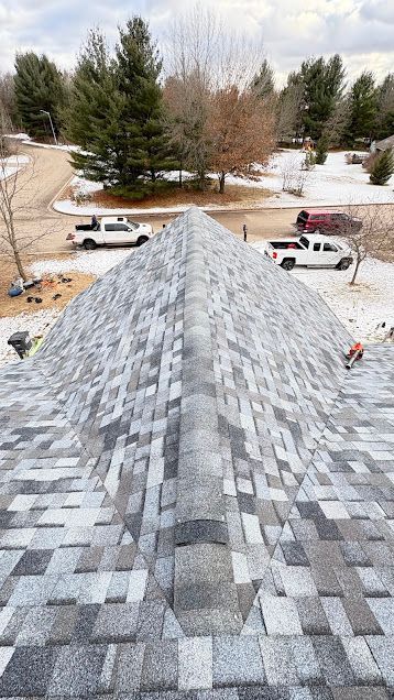 Steep gray shingled roof viewed from the ridge, with snowy yard, trees, and parked trucks below.