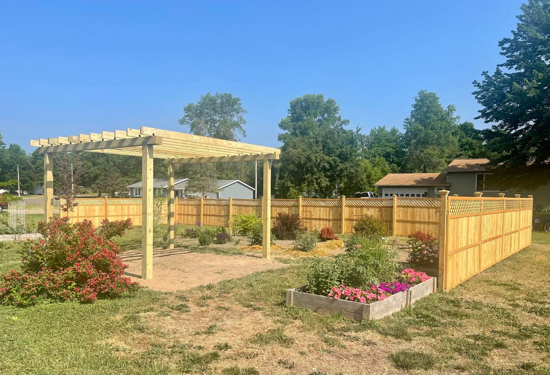 Sunny garden with a wooden pergola, fenced beds, and pink flowers under a clear blue sky