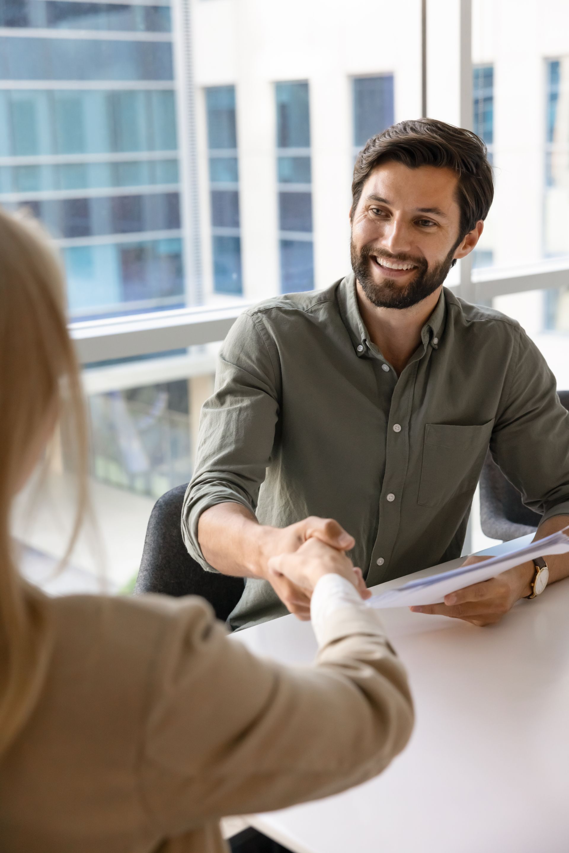 Two people shaking hands across a desk in a bright office, one holding a folder and smiling
