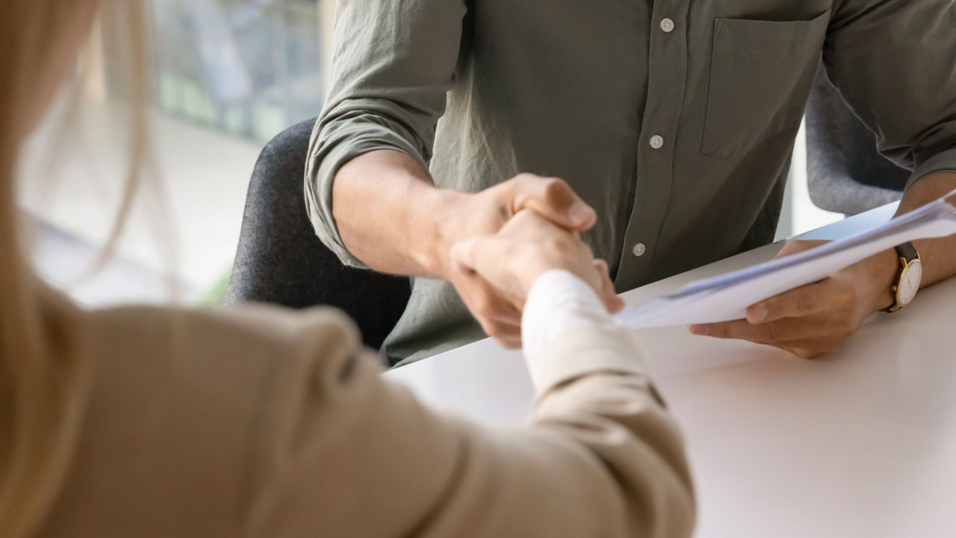 Handshake over documents at a desk, with one person holding a clipboard.