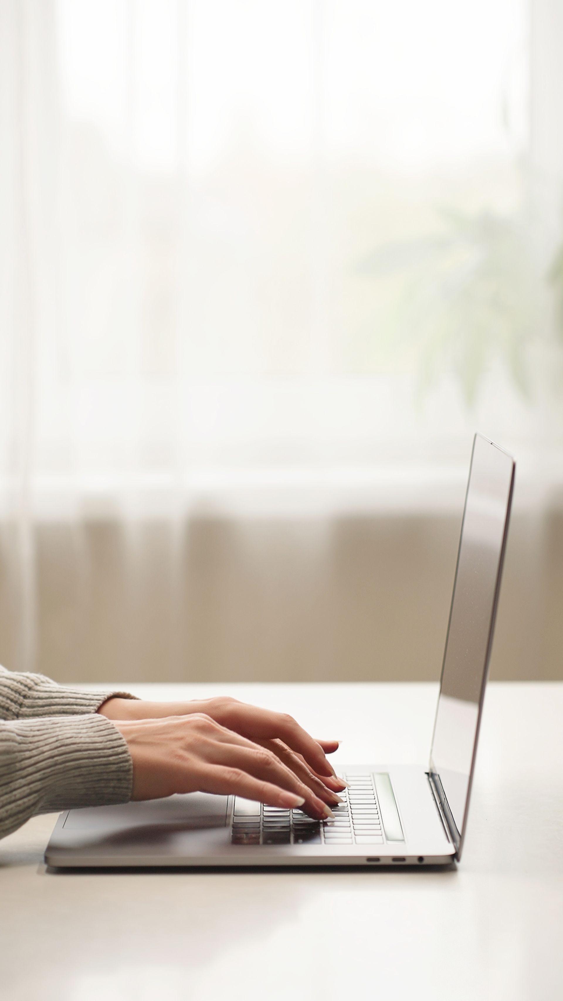 Hands typing on a laptop beside a bright window with sheer curtains