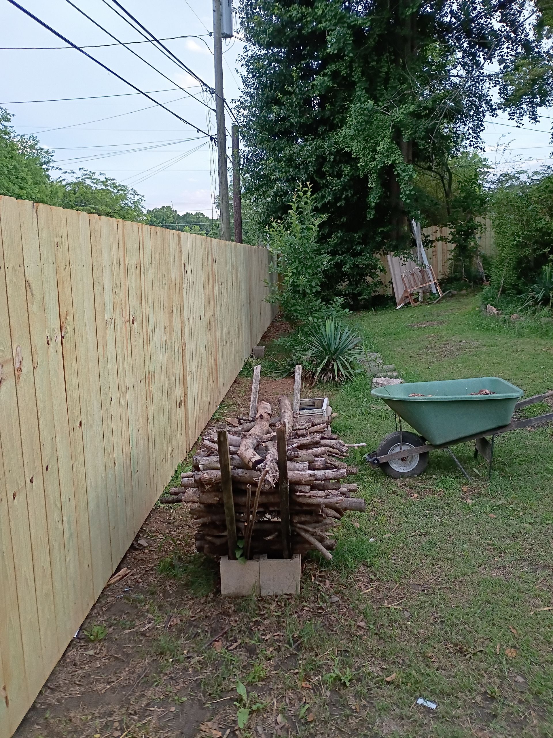 Backyard with a wooden fence, stacked firewood, and a green wheelbarrow on grass