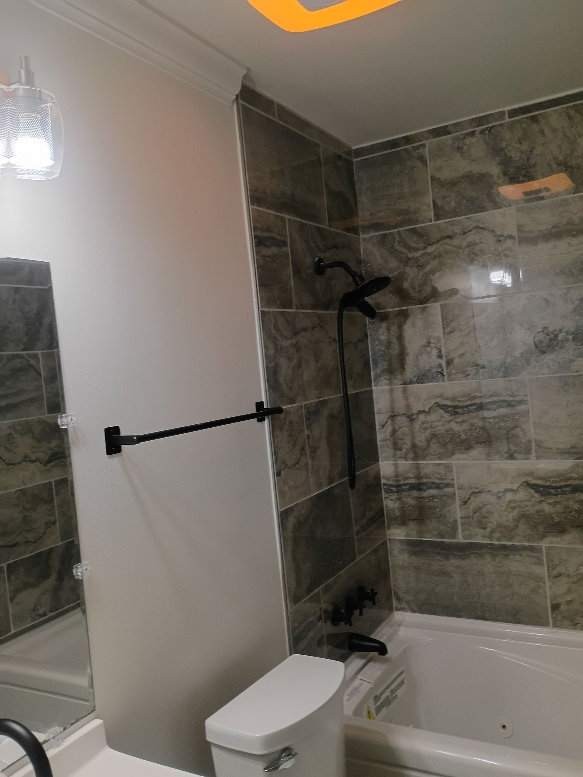 Modern bathroom with white toilet, gray tiled walls, bathtub, showerhead, and black grab bar.
