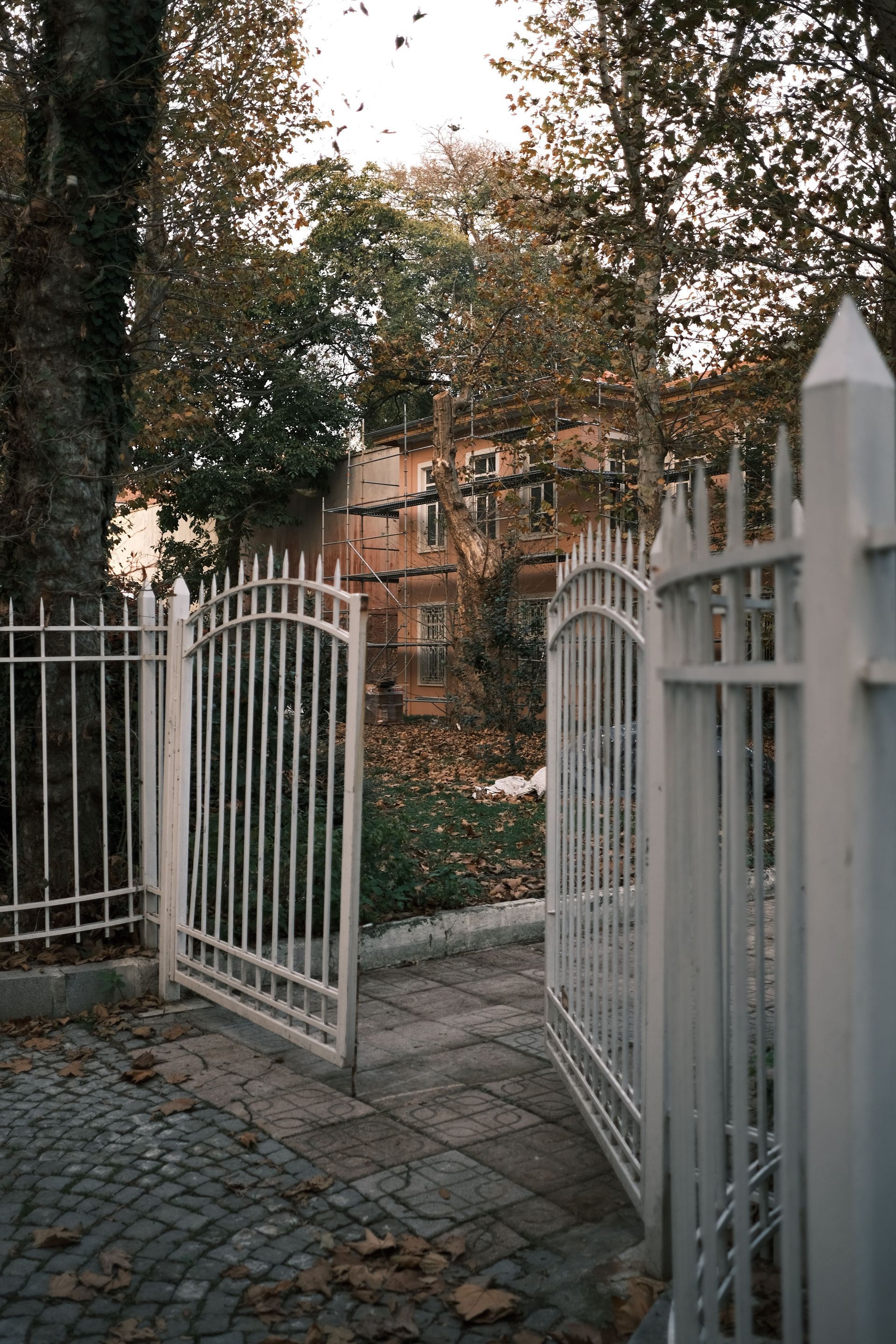 Open white metal gate leading to a brick path and house behind trees
