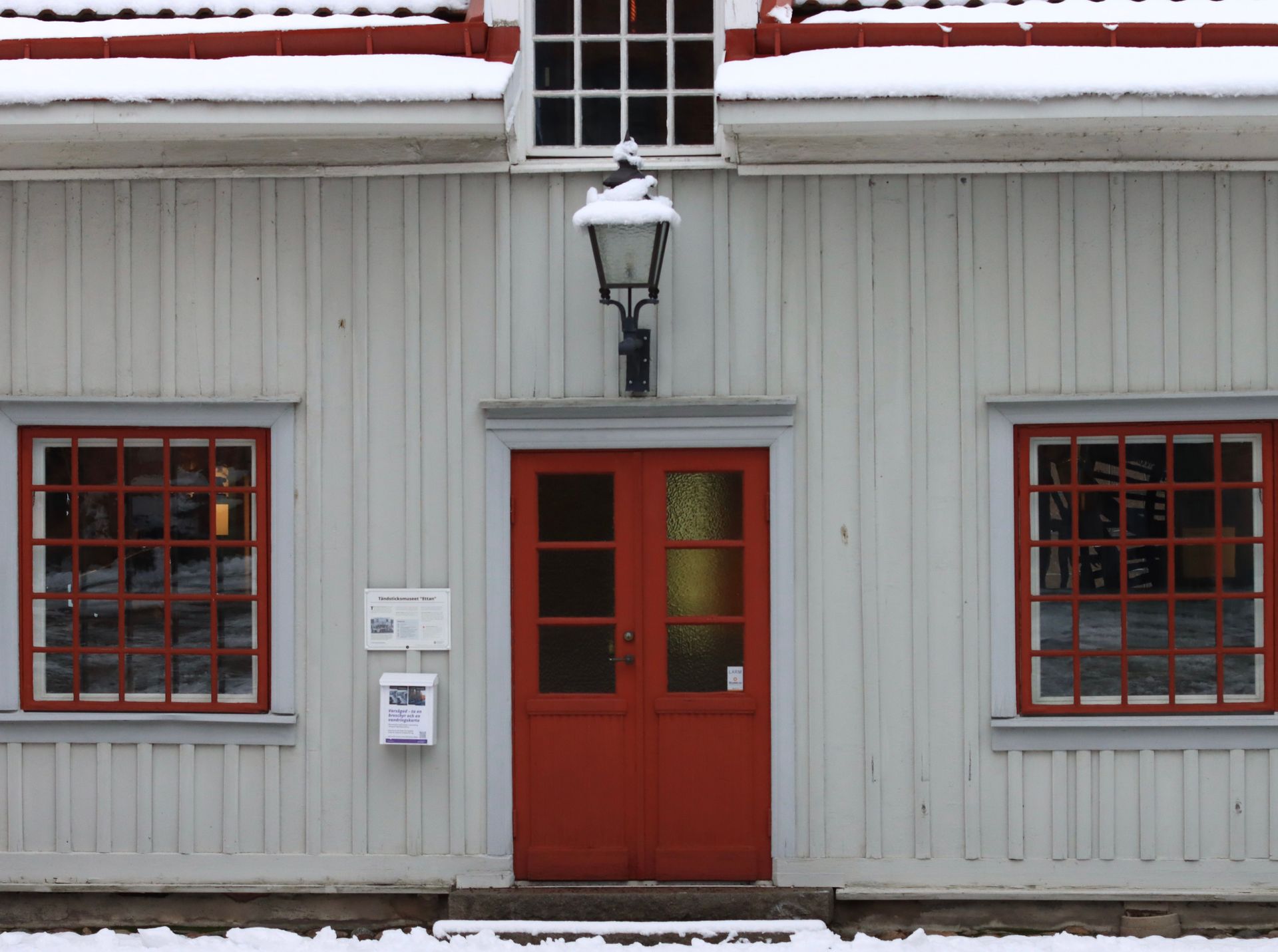 Red door and windows on a gray building with snow on the roof and a wall-mounted lantern above the entrance