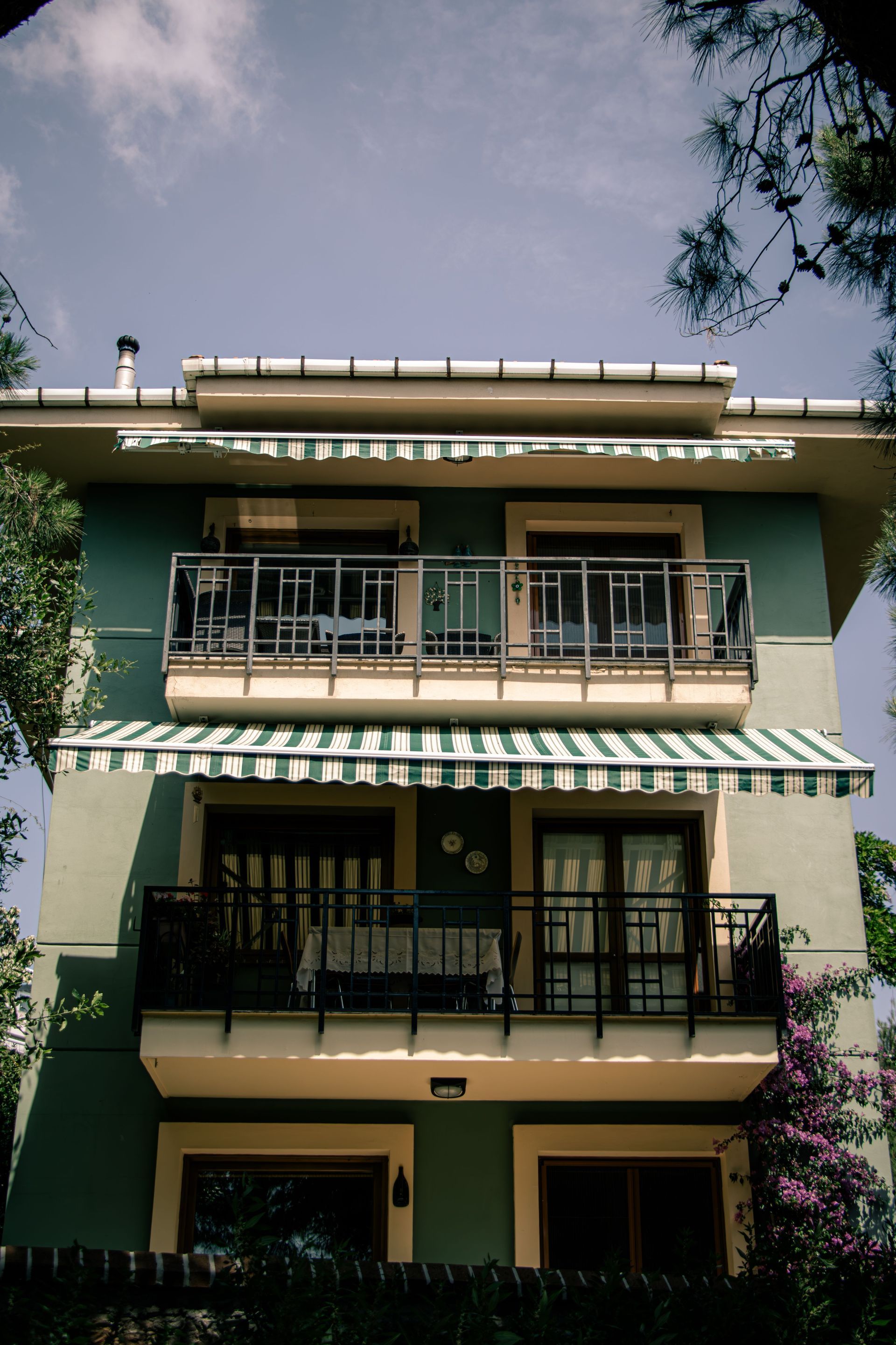 Three-story green apartment building with balconies and striped awnings under a partly cloudy sky