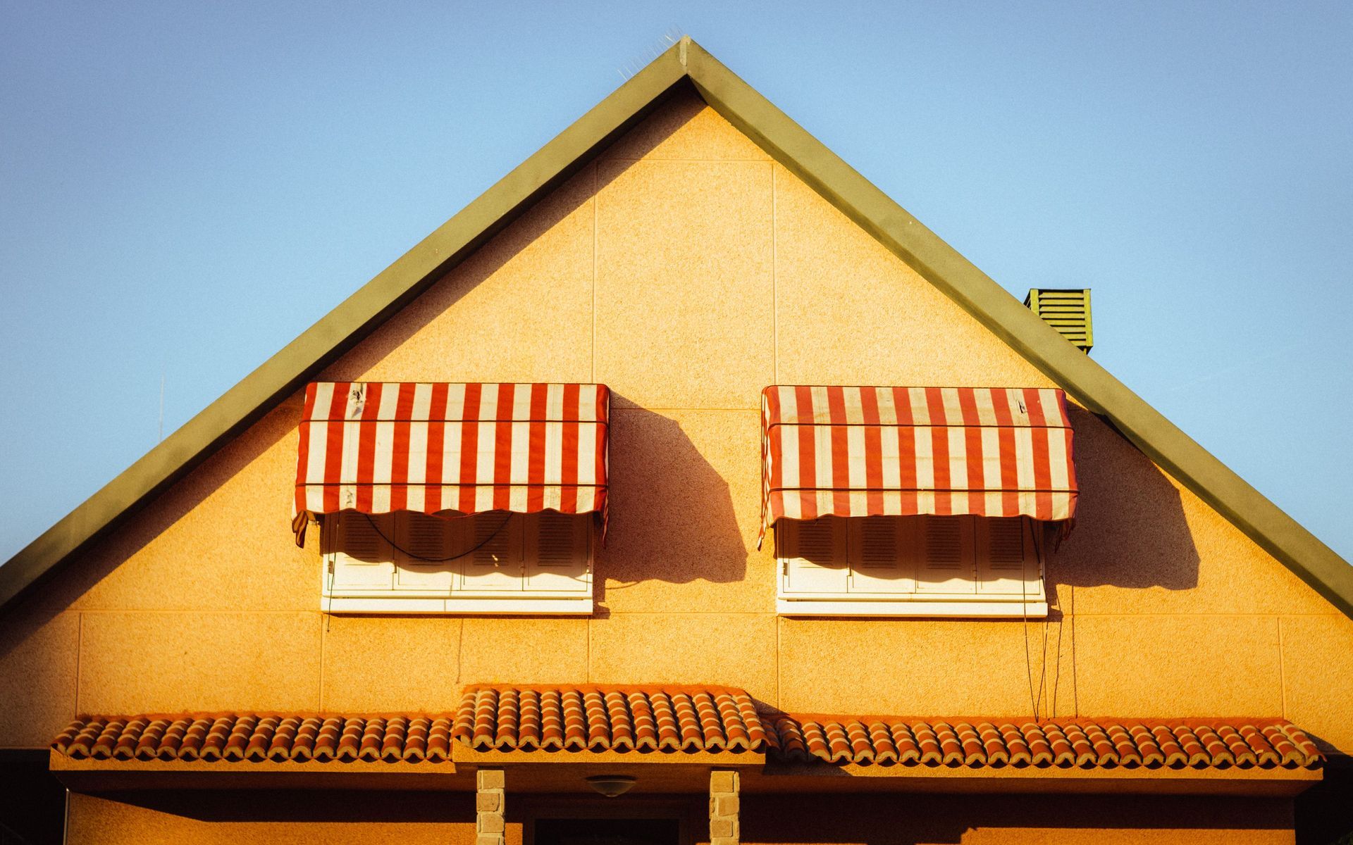 Yellow house wall with two red-and-white striped awnings and terracotta roof tiles against a blue sky